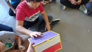 Dzieci siedzą w klasie na podłodze w kole, chłopiec wrzuca kartkę do urny