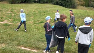 Kilkoro dzieci chodzi po trawie i zbiera śmieci a w tle drzewa