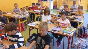 Dzieci siedzą w klasie na krzesełkach przy ławkach, i piszą na kartkach