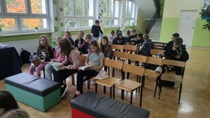 Uczniowie siedzą na pufach i na krzesłach na szkolnym korytarzu