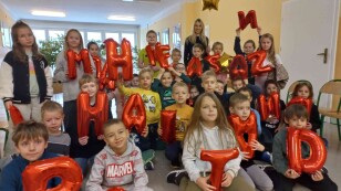 Uczniowie siedzą na krzesełkach i prezentują czerwone balony w kształcie liter