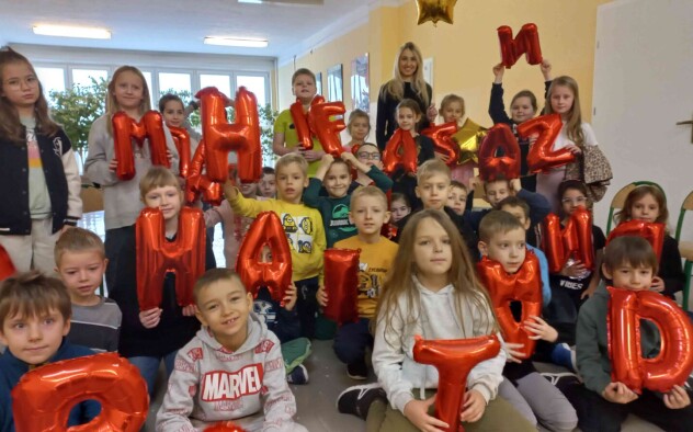 Uczniowie siedzą na krzesełkach i prezentują czerwone balony w kształcie liter