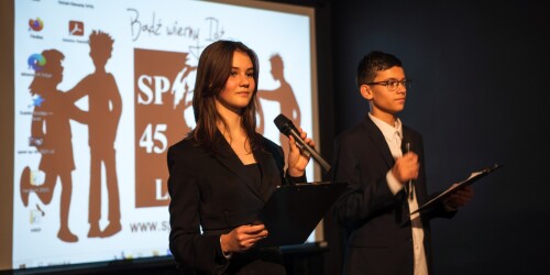 Dwoje uczniów na scenie z mikrofonami na tle logotypu szkoły