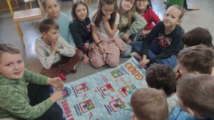 Dzieci czytają z planszy kolejnośc udzielania pierwszej pomocy. Siedzą w kole, w klasie