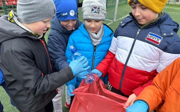 Zdjęcie przedstawia uczniów wrzucających plastikową butelkę do worka na śmieci.
